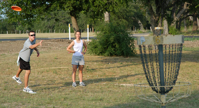 Man and woman standing together as man throws disc into disc golf basket